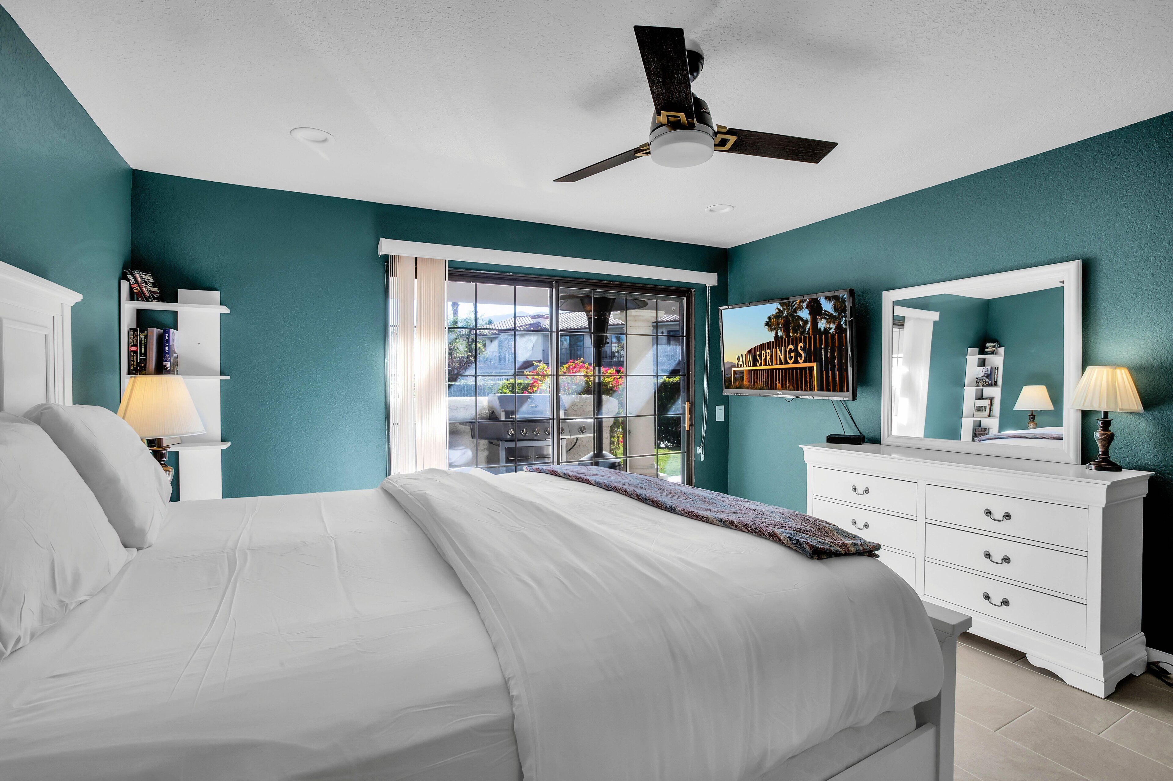 A bedroom with a bed, a ceiling fan, a dresser, a mirror, and a window with a view of a garden.