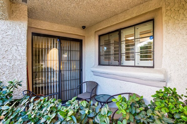 A small outdoor seating area with a table and chairs, a window with metal bars, and potted plants.
