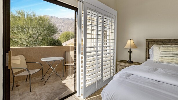A hotel room with a bed, a nightstand with a lamp, a chair, a small table, and a view of the outdoors through a sliding door. - Gated Urban Palm Escape (Palm Springs)