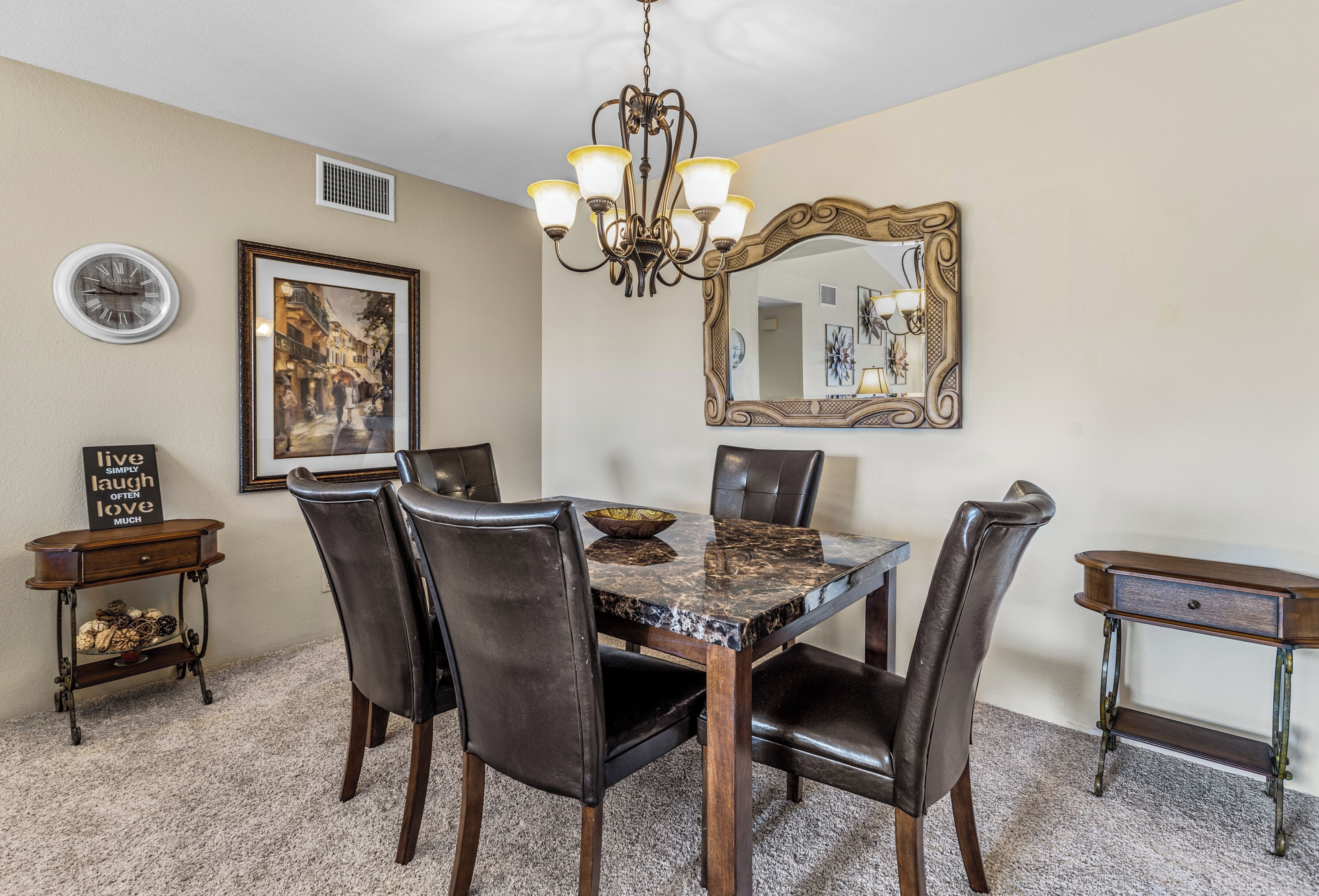 Uma sala de jantar com mesa de mármore, cadeiras de couro, um candelabro, um espelho grande, um relógio e um quadro na parede.