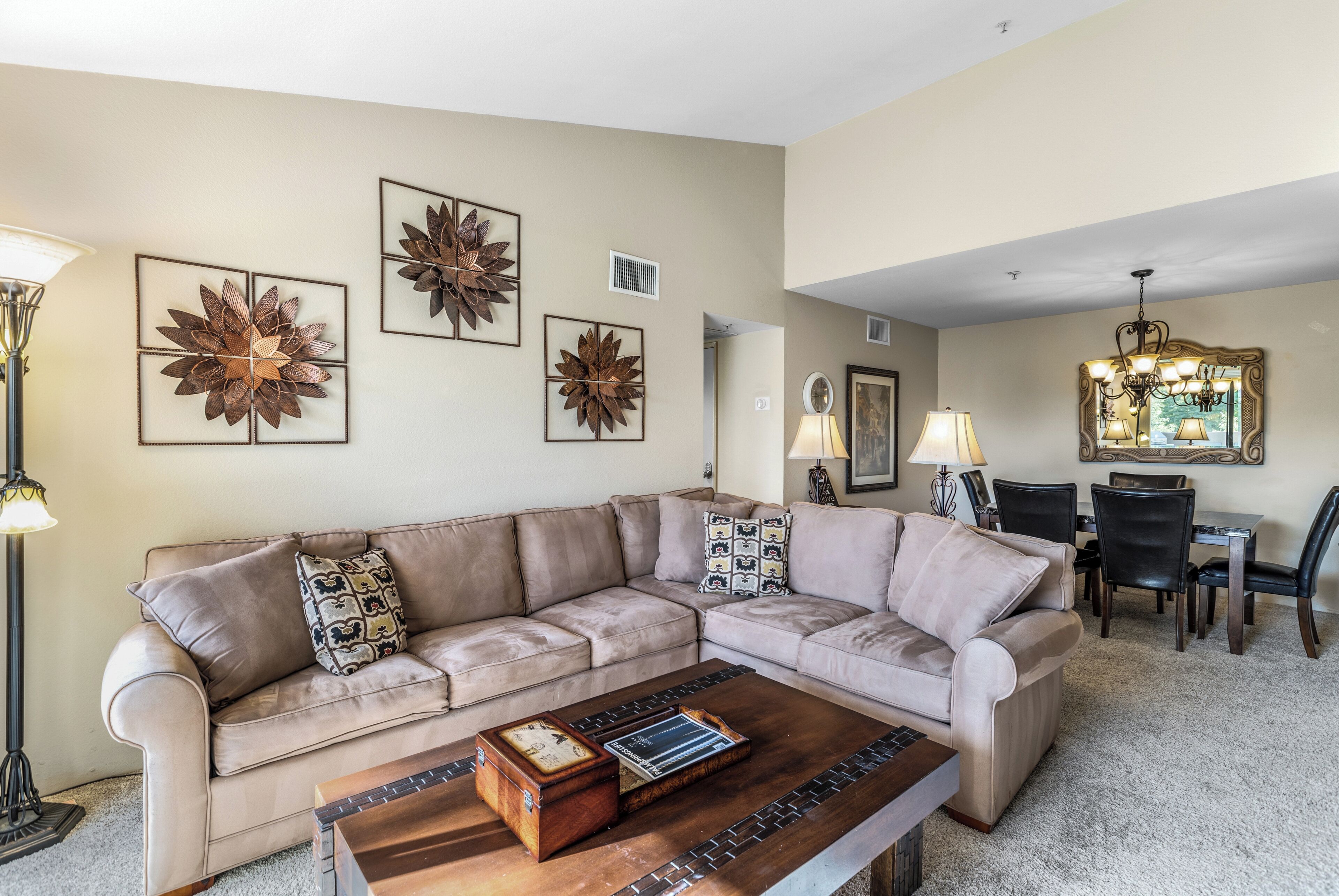 A living room with a beige sectional sofa, a wooden coffee table, and a dining area with a chandelier.