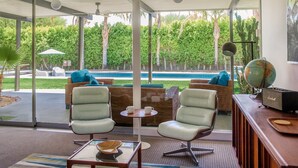 Un salón moderno con puerta de vidrio que da a una zona de piscina, con dos sillones, una mesita y un radio.