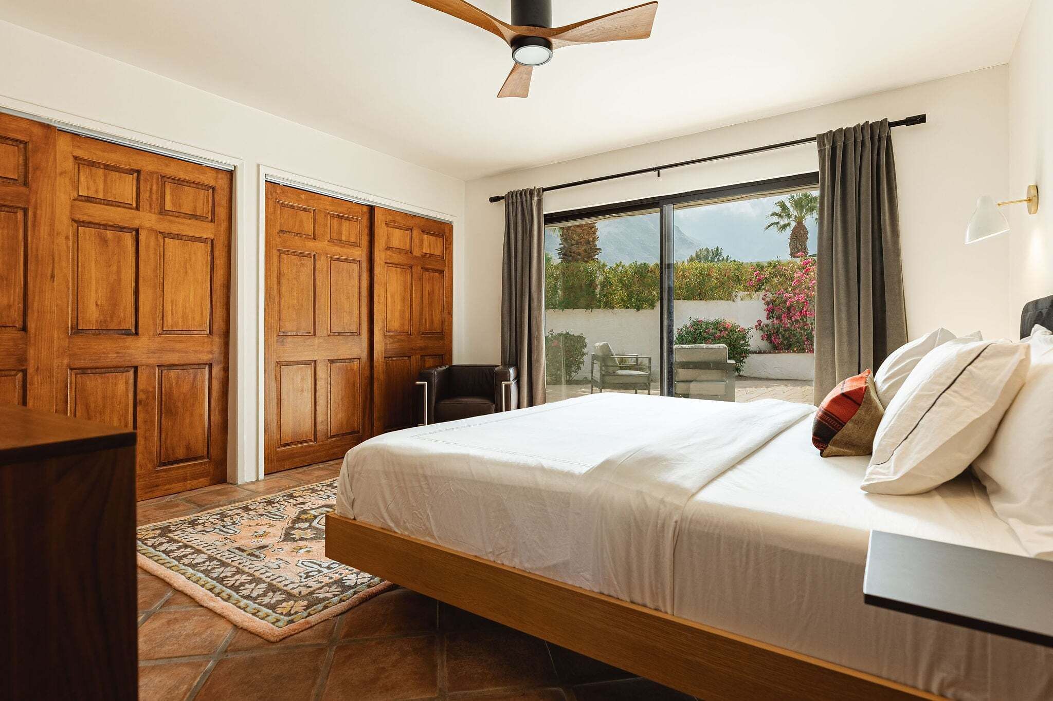 A bedroom with a bed, a ceiling fan, a view through a sliding door to an outdoor area, and a wooden wardrobe.