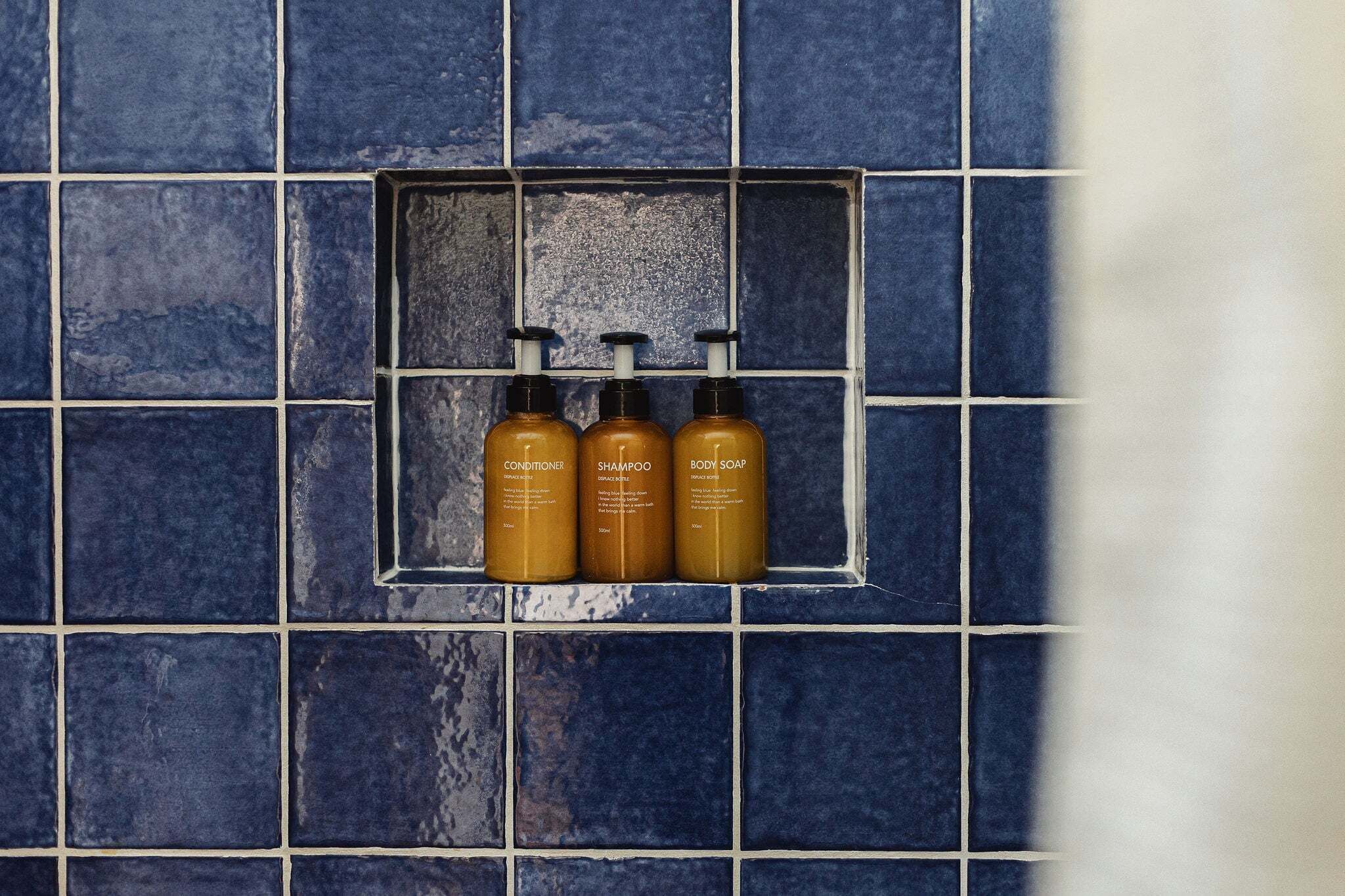 A bathroom with a tiled wall and a shelf holding three bottles of toiletries.