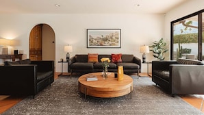 Ein modernes Wohnzimmer mit einem runden Holztisch, schwarzen Sofas, einem großen Gemälde und Blick durch ein großes Fenster.