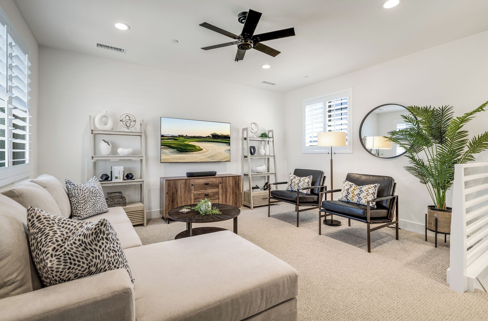 A modern living room with a beige sofa, a coffee table, two leather chairs, a TV, and a ceiling fan.