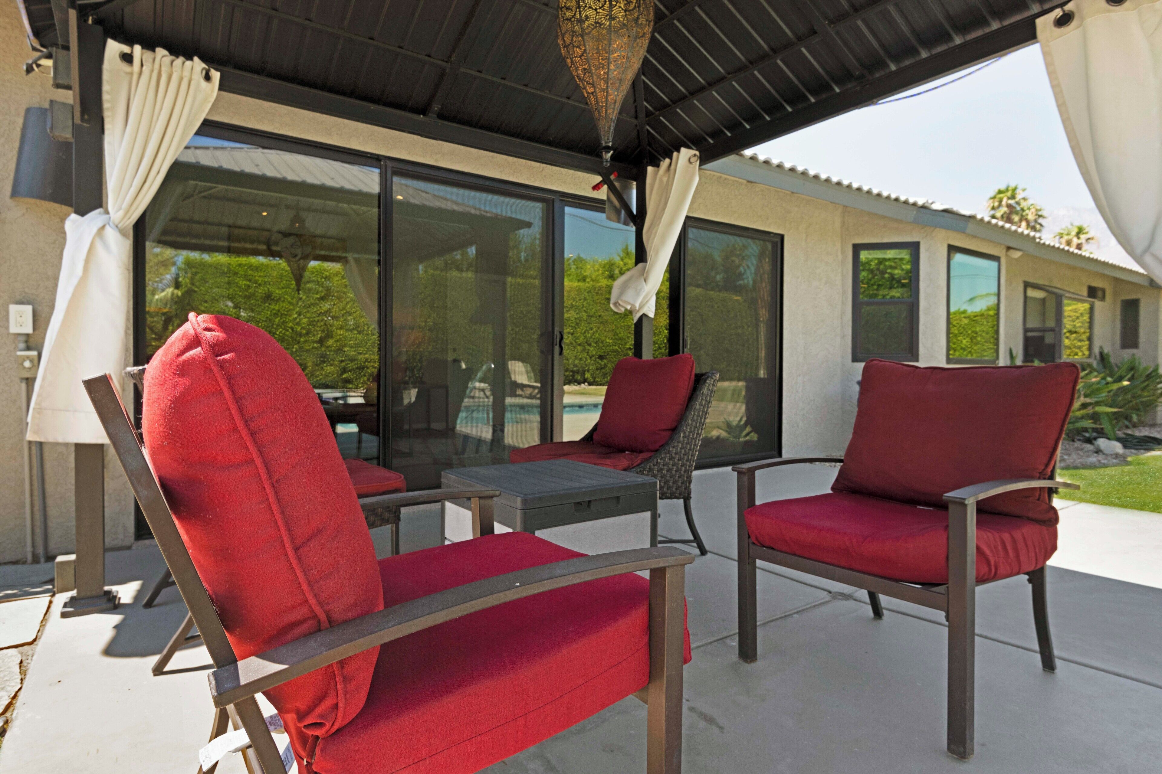 A patio area with two red cushioned chairs and a small table, a glass door leading to a pool, and a covered roof structure.