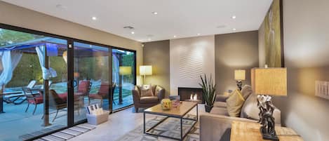 Una sala de estar moderna con puerta de cristal que da a un espacio de estar al aire libre, chimenea y una mesa de centro con objetos decorativos.
