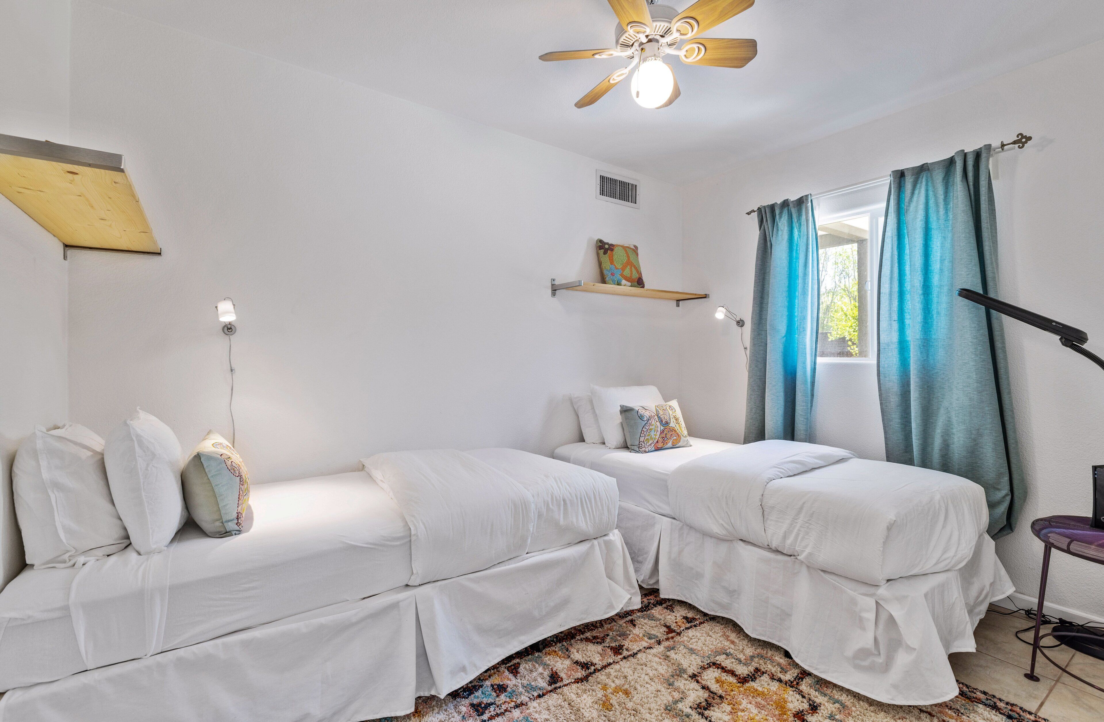 A room with two white beds, a ceiling fan, a window with blue curtains, and a small shelf with a picture.