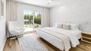 Ein modernes Schlafzimmer mit einem großen Bett, einem weißen Stuhl, einem Deckenventilator und Blick auf Grünflächen.