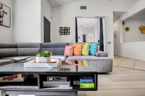 Un salon moderne avec un canapé gris, des coussins colorés, une table basse en verre et une bibliothèque contenant des jeux de société et des livres.