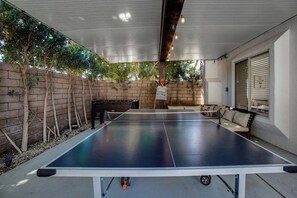 An outdoor ping pong table with a net, surrounded by a brick wall and trees. - Flora Vista Sunshine (Palm Springs)