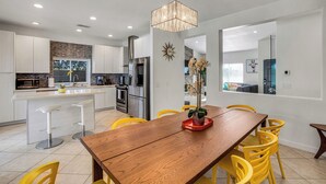 A modern kitchen with white cabinets, stainless steel appliances, and a wooden dining table with yellow chairs. - Flora Vista Sunshine (Palm Springs)