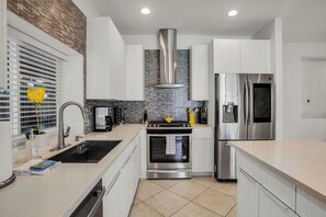 A modern kitchen with white cabinets, stainless steel appliances, a tiled backsplash, and a window with blinds. - Flora Vista Sunshine (Palm Springs)