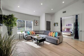 A modern living room with a grey sectional sofa, a coffee table, and a large window with blinds. - Flora Vista Sunshine (Palm Springs)