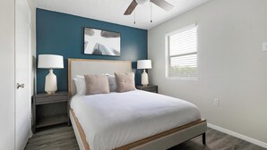 A bedroom with a bed, two bedside tables with lamps, a ceiling fan, a window with blinds, and a wall-mounted artwork.
