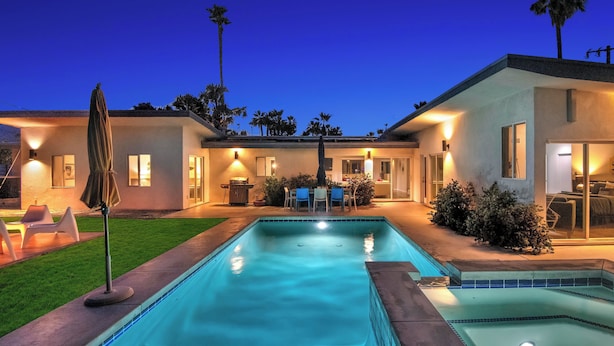 Une piscine avec un espace patio, un coin salon couvert et un bâtiment bien éclairé au design moderne.