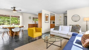 Un salon moderne avec un fauteuil jaune, une table basse en verre, un canapé blanc et un canapé bleu. On y trouve également un coin repas avec une table et des chaises en bois, ainsi qu’une cuisine avec des meubles et un plan de travail en bois.