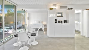 Et moderne, minimalistisk værelse med en skydedør i glas, der fører til et udendørs område, et glasbord med hvide stole, et hvidt skab og et lille tekøkken med kaffemaskine.