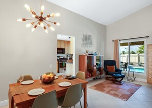 A dining area with a wooden table set for four, a modern chandelier, and a view of the kitchen and pool in the background. - Stylish Boho Treasure (Palm Springs)
