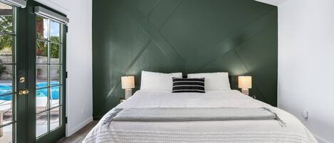 Een slaapkamer met een groene accentmuur, een groot bed met wit beddengoed en een uitzicht op een zwembad door een glazen deur.