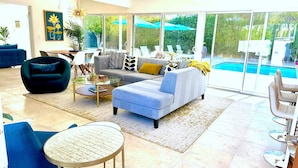 A spacious living area with a sofa, armchair, and round coffee table. A large sliding glass door leads to a pool area. - Serenity in Sunmor (Palm Springs)