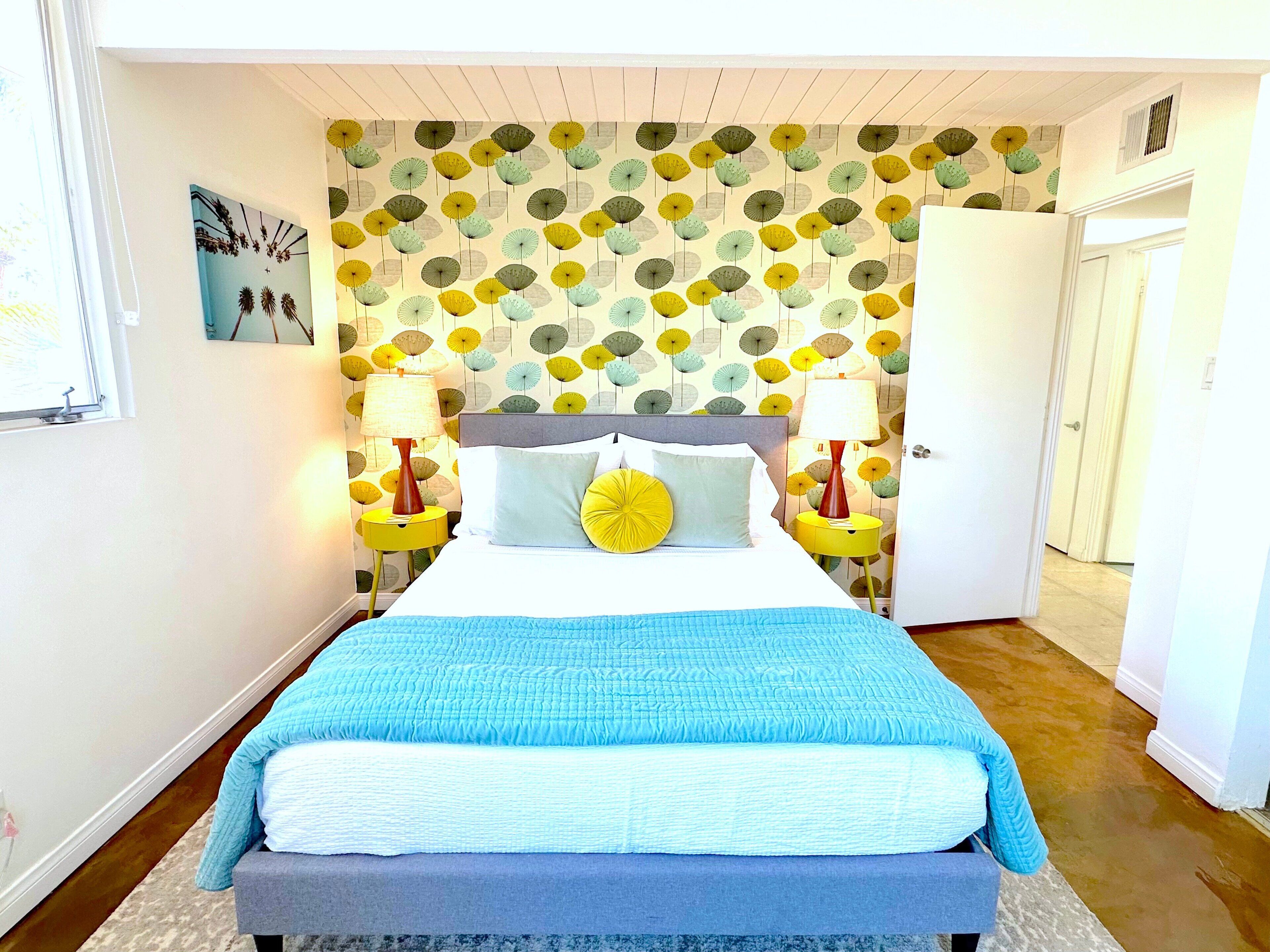 A bedroom with a patterned wallpaper, a bed with a blue quilt, two bedside lamps, and a door to the right.