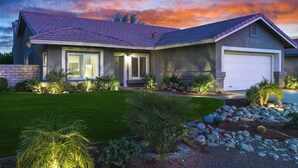 A modern two-story house with a red-tiled roof, a large garage, and a well-maintained front lawn with various plants.