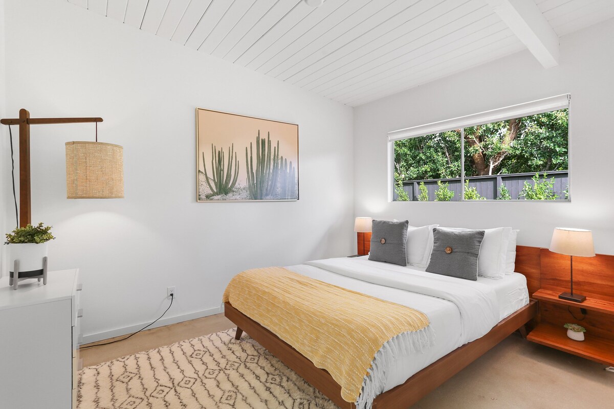 Une chambre moderne avec un lit, des tables de chevet, une lampe suspendue, un tableau accroché au mur et une fenêtre donnant sur un paysage verdoyant.