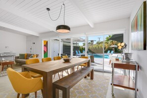 A modern living room with a wooden dining table, yellow chairs, and a bench. There is a pool visible through the glass doors. - Bright & Harmonious Oasis (Palm Springs)