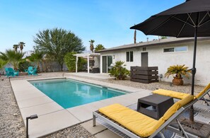 Une piscine entourée de chaises longues et dotée d’un parasol, le tout adossé à un bâtiment blanc agrémenté d’une petite terrasse et de plantes en pot.