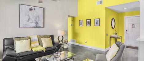 A modern living room with a black leather sofa, a glass coffee table, and a yellow accent wall.