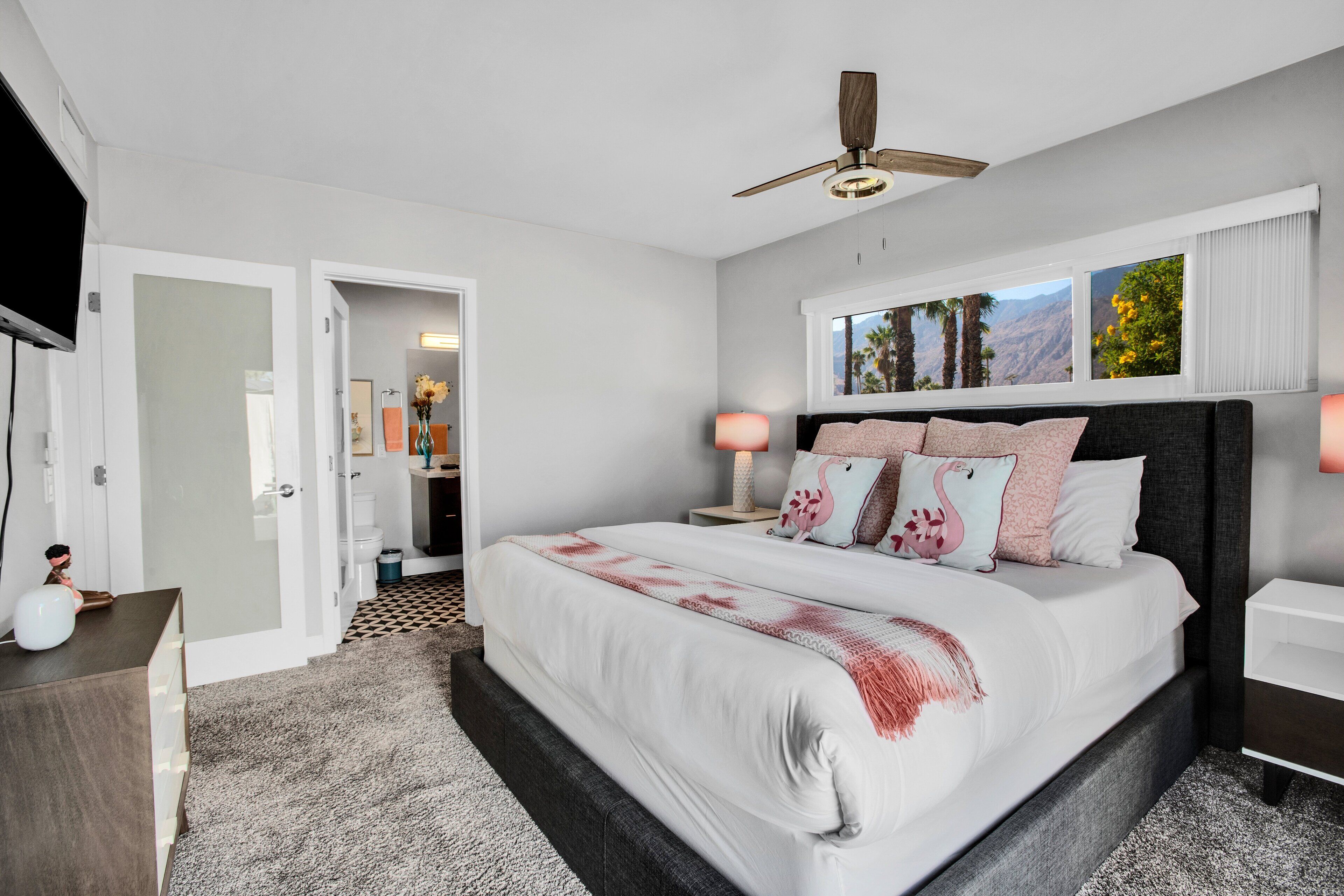 Een moderne slaapkamer met een groot bed, een plafondventilator, een televisie en een raam met uitzicht op palmbomen.