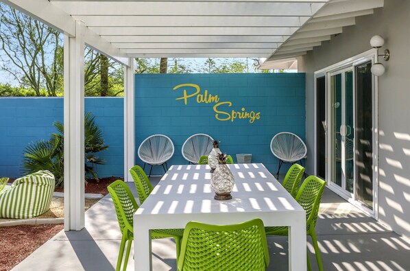An outdoor patio with a white table and green chairs, a blue wall with 'Palm Springs' written on it, and a decorative vase. - Desert Flamingo PS (Palm Springs)