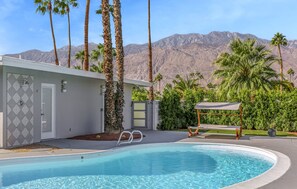 A swimming pool with a mountain backdrop, surrounded by palm trees and a white building with a geometric tile design. - Desert Flamingo PS (Palm Springs)