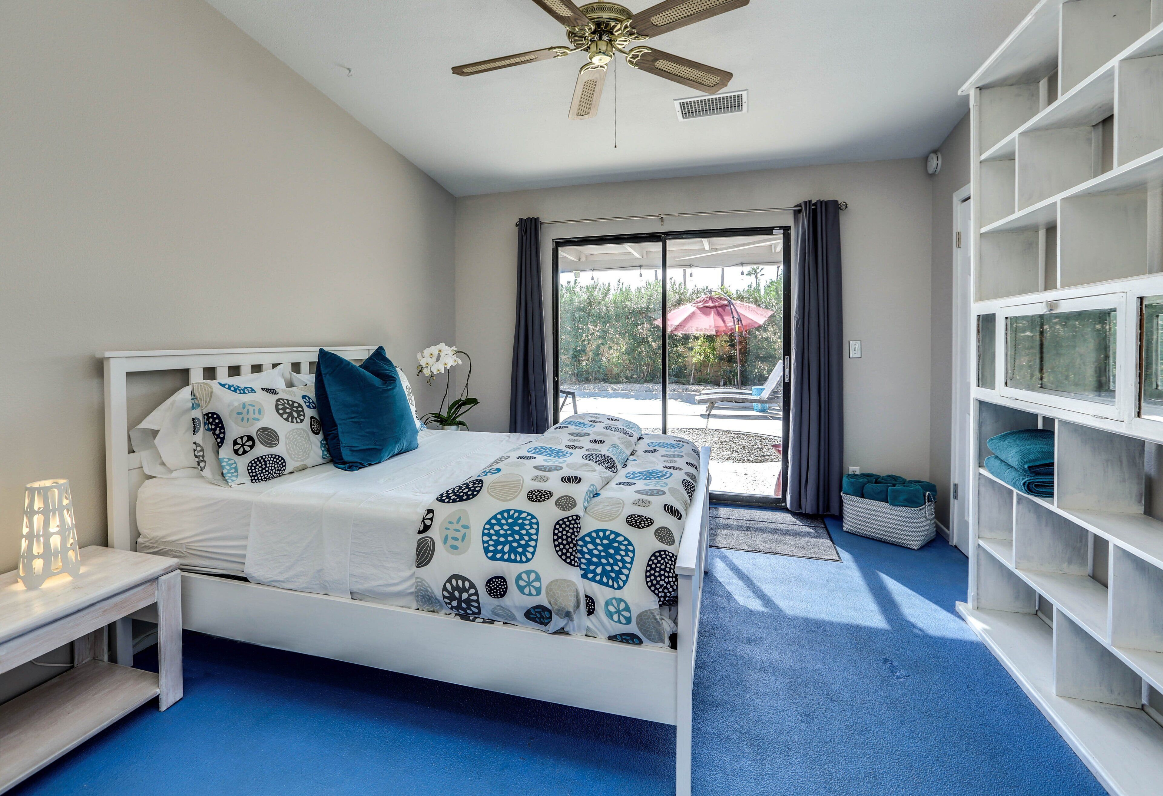 A bedroom with a bed, a nightstand, a ceiling fan, a window with curtains, and a view of the outdoors.