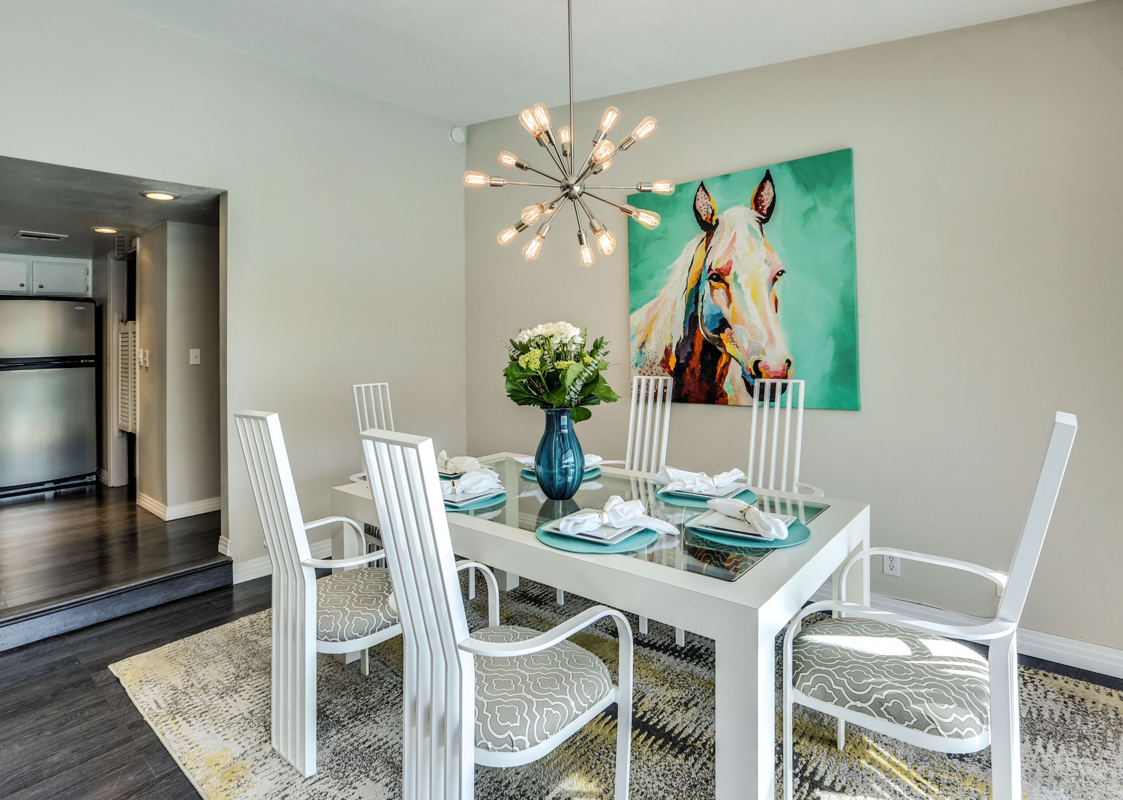 Un comedor moderno con una mesa blanca dispuesta para cuatro personas, rodeada de sillas blancas con cojines estampados. Un jarrón azul con flores está sobre la mesa, y un cuadro abstracto y colorido adorna la pared.