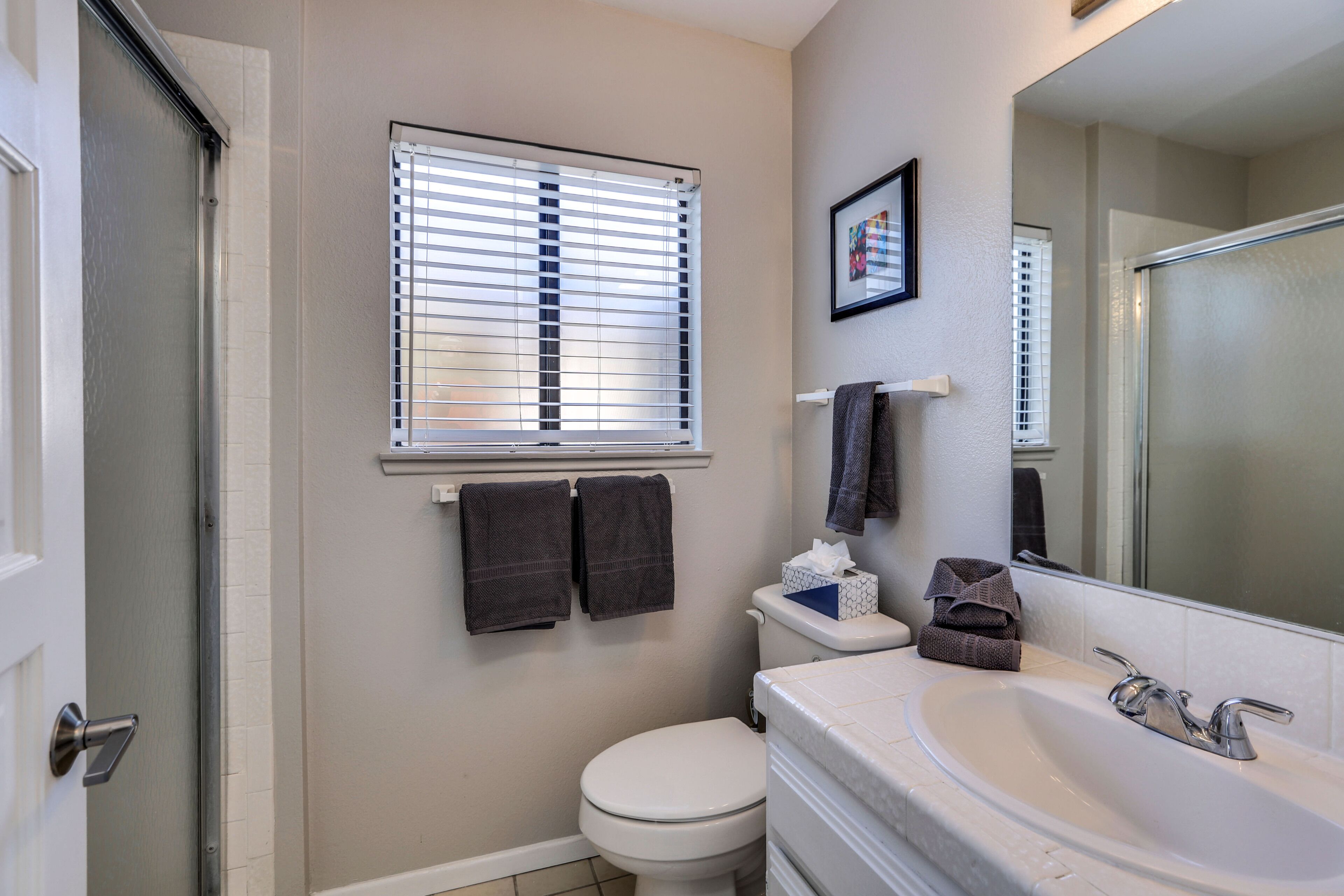Een badkamer met toilet, wastafel en douche. Handdoeken hangen aan de muur en er is een raam met zonwering.