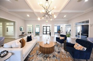 A spacious living room with a modern chandelier, a round wooden coffee table, and a large window offering a view of the gym area. - Landing | Amazing 2BD, Courtyard, Game Room (Murfreesboro)