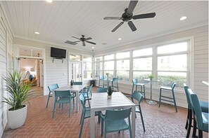 A sunlit room with a brick floor, white walls, and large windows. There are several blue chairs and a table with a potted plant. Two ceiling fans are visible. - Landing | Modern 2BD, Courtyard, Game Room (Murfreesboro)