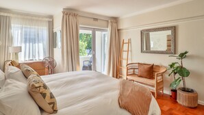 Ein Schlafzimmer mit einem großen Bett, einem Korbstuhl, einer Topfpflanze, einem Spiegel und Blick ins Freie.