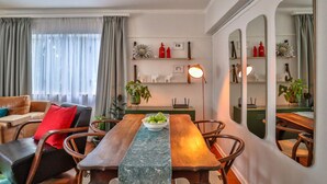Ein Essbereich mit einem Holztisch, Stühlen und einer Schüssel mit Obst. Üchtöpfe und ein Regal mit Dekorationselementen sind ebenfalls vorhanden.