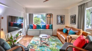 Ein modernes Wohnzimmer mit Fernseher, Deckenventilator, Sofa, Couchtisch und einer Fensterbank mit bunten Kissen.