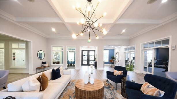 A spacious living room with a modern chandelier, a round wooden coffee table, and a large window offering a view of the gym area. - Landing | Chic 1BD, Game Room, Courtyard (Murfreesboro)
