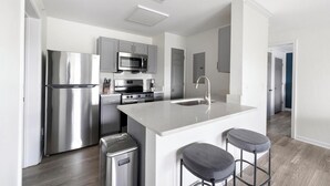 A modern kitchen with stainless steel appliances, a central island with stools, and a view into the adjacent living area. - Landing | Amazing 1BD, Courtyard, Game Room (Murfreesboro)