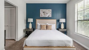 A bedroom with a large bed, two bedside tables with lamps, a blue accent wall, and a window with blinds. - Landing | Spectacular 2BD, Gym (Houston)