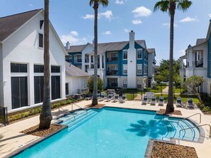 A pool area with lounge chairs, palm trees, and multi-story buildings with balconies. - Landing | Spectacular 2BD, Gym (Houston)