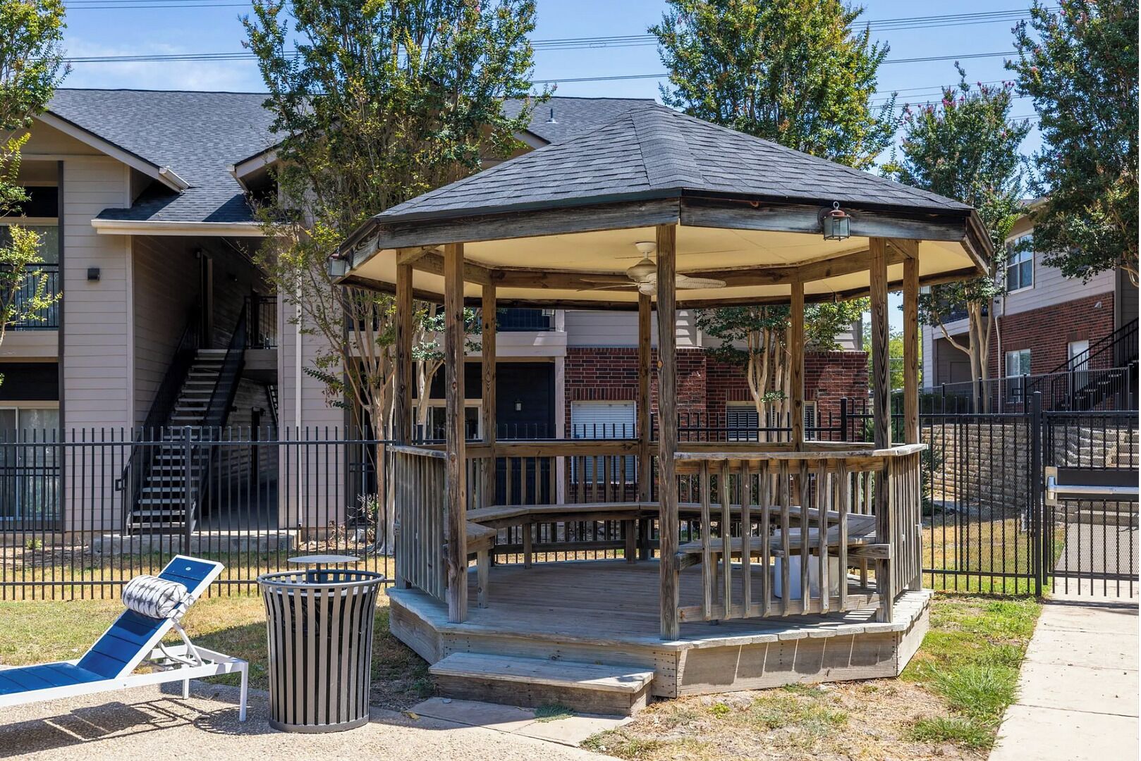 Un kiosque doté d’une structure en bois et d’un toit couvert, entouré d’une clôture, avec une poubelle et un panneau bleu à proximité.