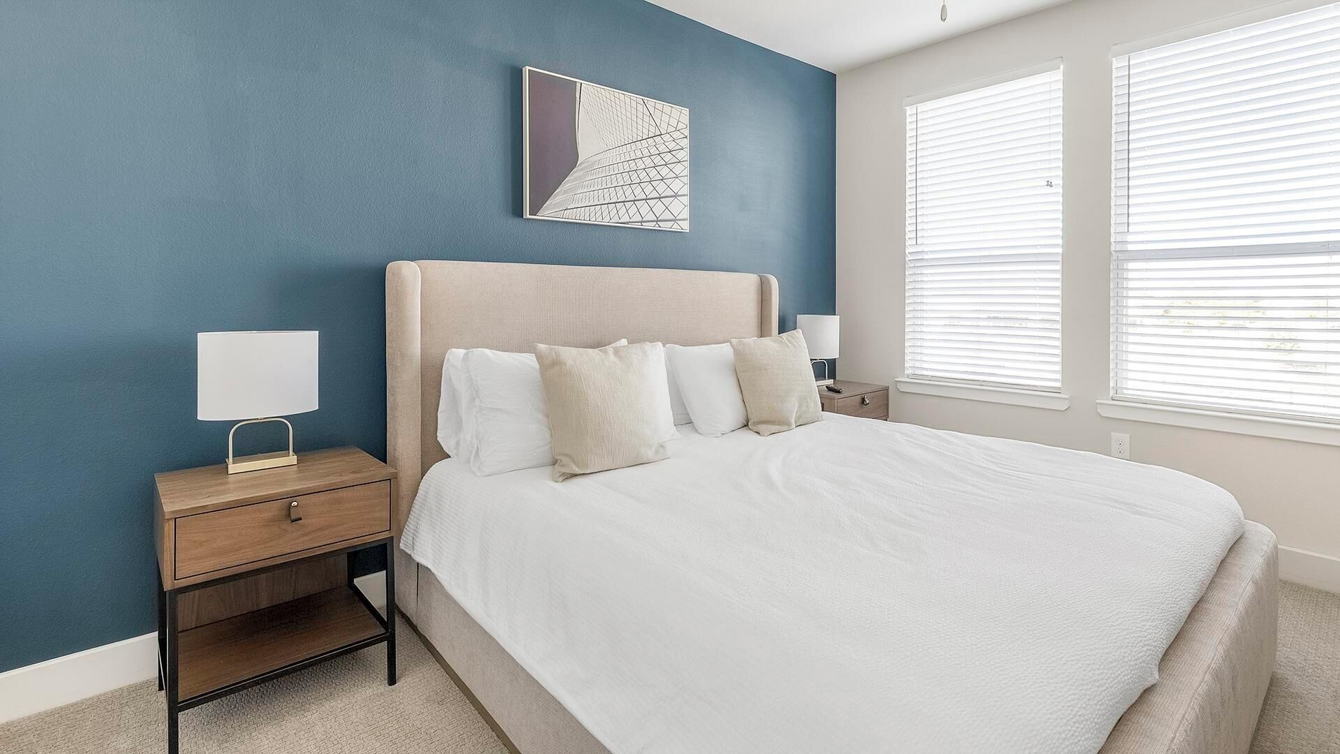 A bedroom with a bed, two bedside tables, a nightstand, a wall art piece, and two windows with blinds.