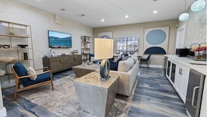 A modern living room with a sofa, coffee table, and a TV. There is a kitchen area with cabinets and a dining table.